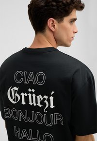 Schwarzes Baumwoll-T-Shirt mit großem weißen Text auf der Rückseite: "CIAO", "Grüezi", "BONJOUR" und "HALLO", in unterschiedlichen Schriftarten.