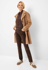 Bruine wollen jas met zwarte knopen, gecombineerd met een donkerbruine shirt en bruine corduroy broek. Lichtbruine sneakers met witte veters.