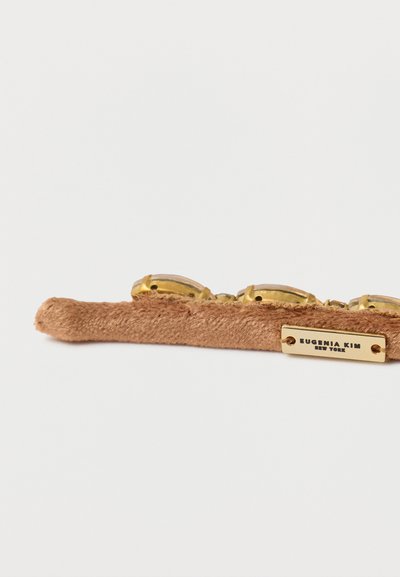 Ruskea mokkainen hiuspanta, jossa on kullanväriset metalliosat, suorakaiteen muotoinen ja EUGENIA KIM NEW YORK -logo painettuna levylle.