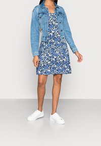 Blaues Blumenkleid mit weißen Blumen, kombiniert mit einer hellen Jeansjacke. Weiße Sneakers vervollständigen das Outfit. Glatter Stoff und lässiges Design.