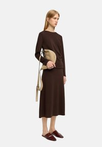 Brązowa bluzka z długim rękawem i midi spódnica z miękkiego materiału. Modelka trzyma jasnobeżową torbę w kształcie półksiężyca na pasku, a na sobie ma czerwone wsuwane buty.