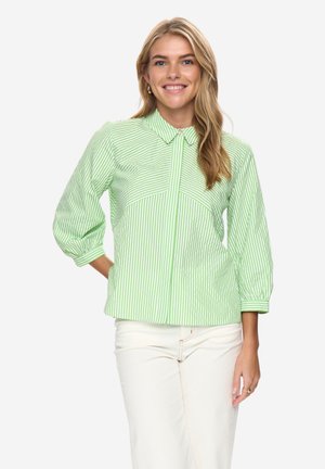 Femme aux longs cheveux blonds portant un chemisier à rayures vertes et blanches avec des manches trois-quarts et un pantalon blanc, souriante, main sur la hanche.