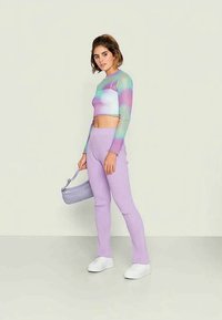 Uma jovem mulher está de pé segurando uma pequena mala de lavanda, vestindo uma t-shirt cropped em tie-dye pastel, calças caneladas lavanda e ténis brancos.