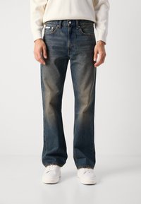 Vaqueros de mezclilla azul con un acabado desgastado, corte recto, cinco bolsillos y cierre de botón, llevados con zapatillas blancas.
