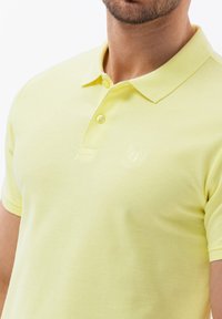 Polo tričko ve světle žluté barvě, s klasickým límcem, dvouknoflíkovou légou a diskrétním vyšitým logem na levé hrudi. Hladká textura.