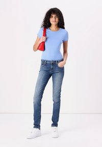 Camiseta ajustada de color azul claro, jeans ajustados azules, bolso rojo y zapatillas blancas. La modelo está de pie contra un fondo sencillo.