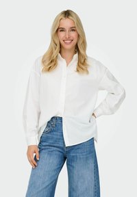 Bijela košulja dugih rukava s gumbima sprijeda, opuštenog kroja, preko plavih širokih traperica. Glatka tkanina s vidljivim detaljima gumba.