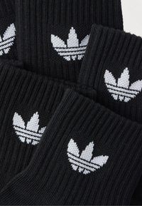 Black ribbed socks featuring a white woven Adidas trefoil logo and three horizontal stripes. Soft texture with a fitted design.