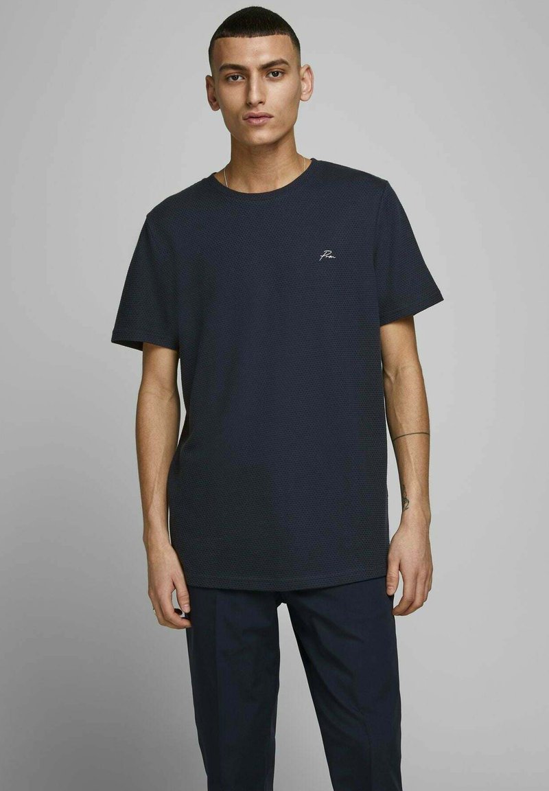 Mørkeblå kortærmet t-shirt med tekstureret stof og et lille hvidt logo på brystet. Sammen med mørke bukser.