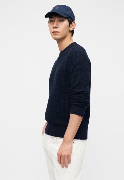 Πλεκτό πουλόβερ ναυτικού μπλε με ριμπ μανσέτες και τελείωμα, σε συνδυασμό με καπέλο ναυτικού μπλε με κόκκινο λογότυπο. Το μοντέλο φοράει εκρού παντελόνια και στέκεται πλαγίως.