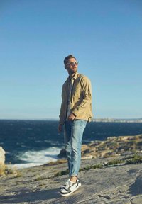 Beige shirtjacket over een grijze shirt, gecombineerd met lichtblauwe jeans en zwart-witte sneakers, staande op ruig terrein bij de zee.