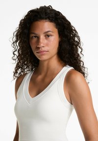 Débardeur blanc côtelé avec un col en V et des bretelles larges, présentant une coupe ajustée qui met en valeur la silhouette et une texture lisse.