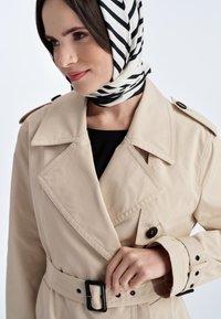Beżowy płaszcz trencz z dużym kołnierzem, podwójnym zapięciem i czarnym paskiem. Czarne guziki oraz teksturowany, w paski czarno-biały szal.