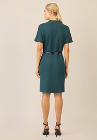 Vestido de punto en color teal con mangas cortas, cintura ajustada y un cinturón negro. Presenta una silueta recta y una textura suave, combinado con sandalias de tacón alto.