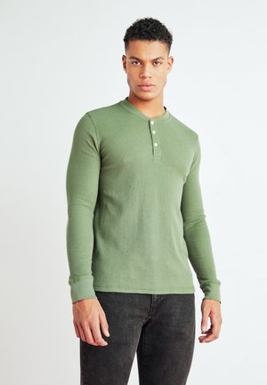 Olivengrøn ribbet langærmet skjorte med henley-udskæring, tre knapdetaljer og tætsiddende manchetter. Sammen med mørke jeans.