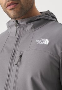 Grijze zip-up jack met een capuchon, voorzien van een logo op de linkerborst, textuurstof en een ritszak aan de voorkant.