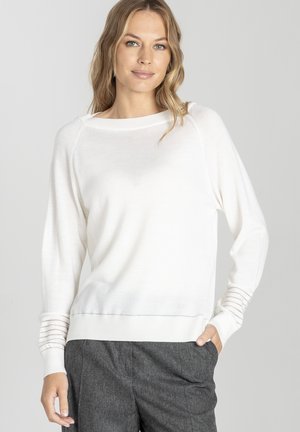 Kobieta z falującymi blond włosami, ubrana w białą sweter z długim rękawem i przezroczystymi, paskowanymi mankietami oraz ciemnoszare spodnie, stoi z jedną ręką w kieszeni.