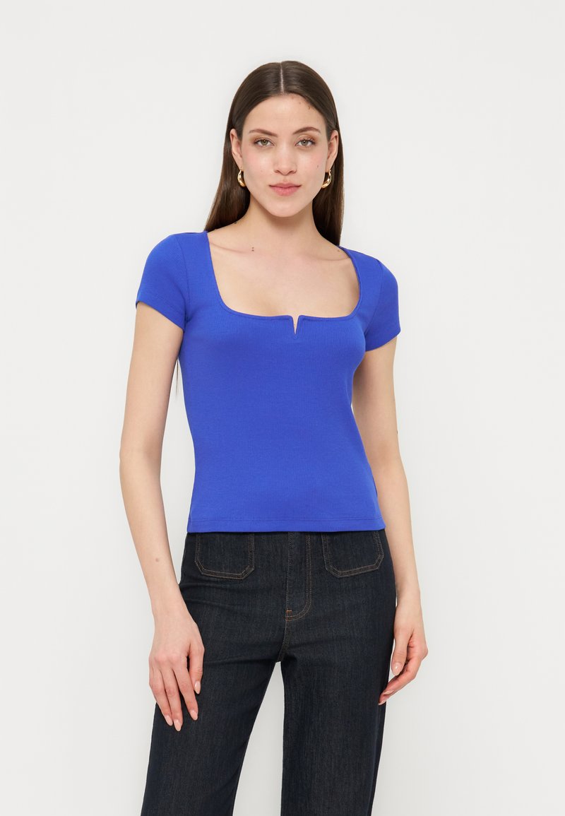 Νέα γυναίκα με μακριά καστανά μαλλιά που φορά χρυσούς κρίκους στα αυτιά, μπλε κοντομάνικη μπλούζα με τετράγωνη λαιμόκοψη και σκούρο τζιν ψηλόμεσο παντελόνι.