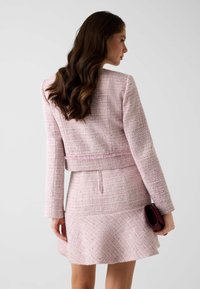 Ensemble veste et jupe en tweed rose pâle avec tissu texturé et motifs subtils. La jupe présente un ourlet évasé ; tenant une pochette sombre.
