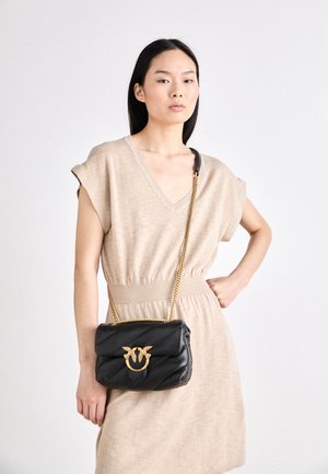 Donna che indossa un abito beige con maniche a copri-spalla e una borsa nera trapuntata con catena dorata e chiusura a forma di uccello sulla spalla.