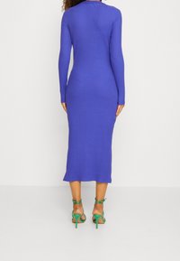 Randig, långärmad klänning i livlig blå färg, midi-längd med en figurnära silhuett. Kombinerad med gröna högklackade sandaler med remmar.