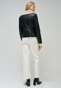 Giacca di pelle nera con colletto beige in pelliccia, abbinata a pantaloni in denim bianchi. I dettagli includono un design aderente e una leggera differenza di consistenza.