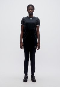 Ενήλικας που φοράει μαύρα αθλητικά ρούχα και μαύρα παπούτσια με πέλματα, στέκεται όρθιος μπροστά σε απλό λευκό φόντο.