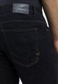 Les jeans en denim bleu foncé présentent des coutures contrastées, une patch en cuir avec le logo et un design distinctif des poches arrière avec des détails uniques.