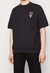 Svart sweatshirt med korta raglanärmar, ribbade muddarna och rundad halsringning. Har en röd och vit broderad grafik på bröstet.