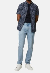 Mann trägt weiße Sneakers, hellblaue Jeans, ein schwarzes T-Shirt und ein unknöpftes kurzärmliges marineblaues Hemd mit Blättermuster, steht mit den Händen hinter dem Rücken.