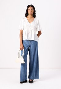 Blusa bianca ricamata con scollo a V e maniche corte a palloncino. Abbinata a jeans a vita alta e gamba larga a righe blu. Tiene in mano una piccola borsa bianca.