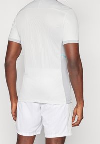 Pánské svetlo šedé sportovní tričko s síťovanou texturou, krátkými rukávy a jemným tyrkysovým akcentem na straně. V kombinaci s bílými šortkami.