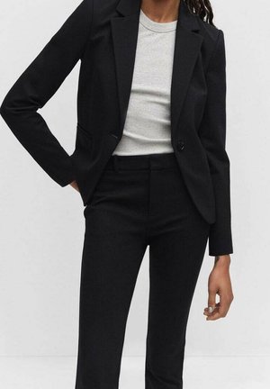 Person trägt einen taillierten schwarzen Blazer und passende schwarze Hose mit einem hellgrauen Rundhals-Shirt und steht mit einer Hand in der Tasche.