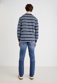 Camisa de manga larga a rayas en navy, azul claro y blanco. Corte holgado, puños acanalados. Llevada con jeans azul desgastados y zapatos de color claro.
