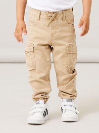 Beige Cargo-Hosen mit elastischen Bündchen, seitlichen Taschen und einem verstellbaren Bund, kombiniert mit weißen Sneakers mit schwarzen Streifen.