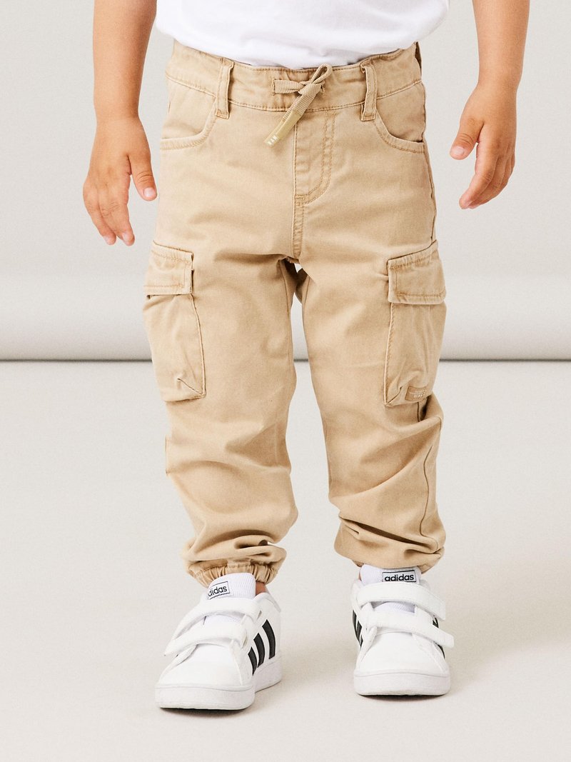 Beige Cargo-Hosen mit elastischen Bündchen, seitlichen Taschen und einem verstellbaren Bund, kombiniert mit weißen Sneakers mit schwarzen Streifen.