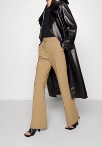 Svart läder trench coat över en svart topp, tillsammans med beige vida byxor och svarta öppna klackar. Släta texturer och skräddarsydd design.