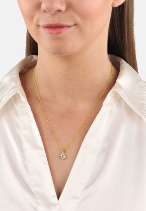 Collana in oro con pendente a forma di gemma rotonda e trasparente. La catena ha un design delicato e intrecciato, indossata sopra una blusa di satin chiaro.
