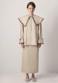 Béžová blůza s oversized volánkovým límcem, dlouhými rukávy s nařasenými manžetami, spárovaná s odpovídající dlouhou sukní. Woven béžové nazouvací boty.