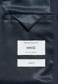 Tissu de costume bleu marine avec une texture lisse ; les étiquettes indiquent "LA COLLECTION DE COSTUMES", "MNG", et "SLIM FIT", indiquant une taille EUR 50, UK 40.