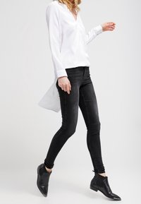 Blusa blanca de manga larga con cuello en V y un dobladillo curvado, combinada con jeans negros ajustados y botas negras hasta el tobillo. Tela suave y corte entallado.