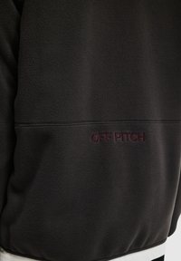 Zwarte fleece sweater met rode geborduurde tekst "OFF PITCH", gemaakt van textured stof en met een gestroomlijnd, minimalistich ontwerp.