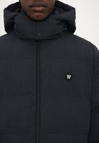 Padded black jacket featuring a grid pattern, high collar, and zip closure. Includes a logo patch and snap buttons on the hood.