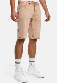 Shorts en denim brun clair à coupe droite, longueur mi-cuisse, avec une texture subtile et des détails de couture, associés à des baskets blanches et des chaussettes.