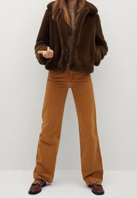 Brązowa kurtka z sztucznego futra z kołnierzem, noszona na szarym topie; zestawiona z żółto-brązowymi spodniami dzwonami i brązowymi mokasynami z akcentem w postaci łańcuszka.