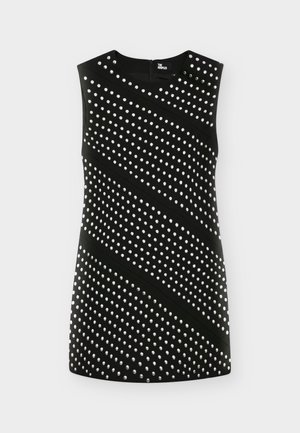 Femme portant une mini robe noire sans manches avec des motifs cloutés en diagonale et des bottes en cuir noires mi-mollet, debout devant un fond uni.