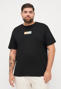Crna majica kratkih rukava od pamuka, s pravokutnim logotipom "JACK & JONES" u narančastoj i tirkiznoj boji na prednjoj strani.