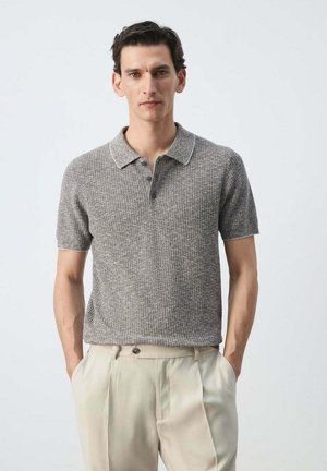 Hombre con camisa polo de punto gris de manga corta y pantalones beige de tiro alto, de pie con las manos en los bolsillos contra un fondo liso.