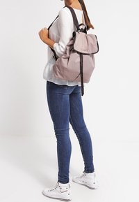Zaino grigio chiaro con chiusura a coulisse, dettagli neri e spallacci regolabili, portato da una persona che indossa una blusa bianca e jeans.