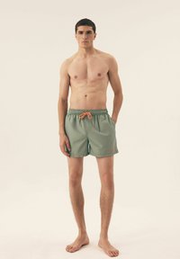 Zielone kąpielówki z elastycznym paskiem w talii i pomarańczowym sznurkiem, z jedną boczną kieszenią i prostym wzorem.